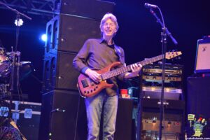Bass guitarist playing on stage, smiling, with large speaker cabinets behind him.