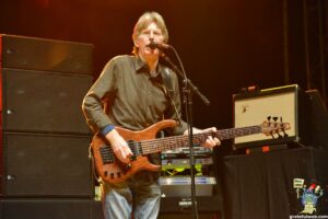 Male guitarist on stage playing a wooden double‑neck bass, singing into a microphone with large amplifiers behind him