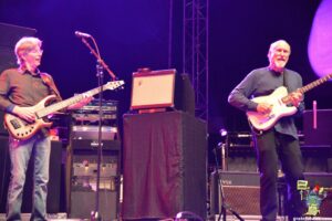 Two older men playing electric guitars on a stage during a live concert, with amplifiers and purple lighting in the background.