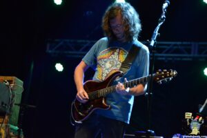 Male guitarist with long hair and a blue graphic t-shirt, playing a sunburst electric guitar on stage. Notable stage lighting in blue hues visible behind him, microphone stand nearby.