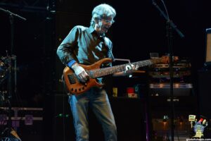 Male guitarist playing a wooden bass guitar on a dimly lit stage with amplifiers and microphones around him