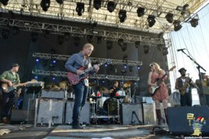 Band performing on an outdoor stage with multiple guitarists and a drummer, under rigging and stage lights.