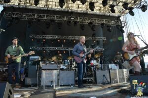 Band performs live on an outdoor stage with guitars and drums under overhead truss and lights.