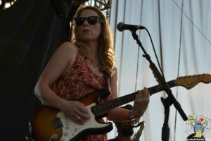 Female guitarist wearing sunglasses and a patterned sleeveless top performing on stage with a Fender Stratocaster, microphone nearby.