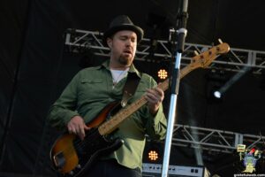 Male musician playing a sunburst bass guitar on stage, wearing a green shirt and fedora hat, focused on his performance.