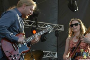 Two guitarists perform on a stage: a man with long blond hair in a blue shirt playing a red electric guitar, and a woman wearing sunglasses playing a guitar with stickers on a patterned dress.