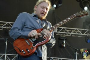 Male guitarist on a stage playing a red electric guitar, wearing a blue shirt, focused on his strings with stage rigging in the background.