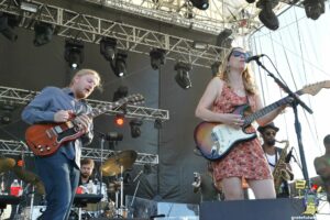 A female guitarist sings into a microphone on stage while another guitarist plays beside her, with a drummer and stage rigging in the background on a sunny outdoor concert.