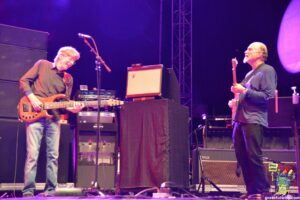 Two musicians on a dimly lit stage playing electric guitars, with amplifiers and equipment in the background under purple lights.