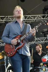 Guitarist playing a red electric guitar on stage, wearing a blue shirt with stage rigging behind him.