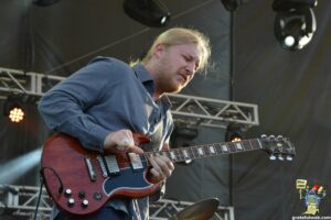 Male guitarist playing a red electric guitar on an outdoor stage, wearing a blue shirt, with stage lighting and rigging in the background.