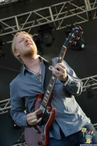 Male guitarist with long blond hair and beard playing a red Gibson electric guitar on stage.