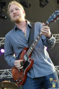 Male guitarist on stage playing a red Gibson-style electric guitar during a live performance on stage lights in the background.
