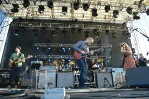 Band performing on an outdoor concert stage with guitarists center, bassist left, singer right, and drums behind them under a metal truss rig