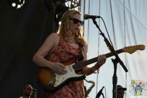 Female guitarist wearing sunglasses, playing a sunburst Fender Stratocaster on stage with a microphone nearby.
