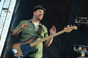 Smiling bass guitarist in a green shirt and black hat playing a sunburst Fender bass on stage at an outdoor concert.