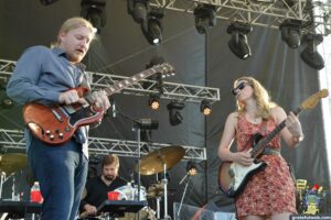 Two musicians perform on an outdoor stage: a man with a red electric guitar on the left and a woman playing a bass on the right.