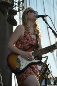 Female guitarist singing into a microphone on stage, wearing sunglasses and a red patterned dress with a sunburst electric guitar