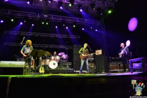 Band performs on a lit stage with purple and green lights, guitarist left, bassist center, drummer in back under metal truss rigging.