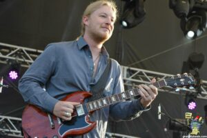 Male guitarist with blond hair in a blue shirt playing a red electric guitar on a stage with rigging and purple lights in the background.