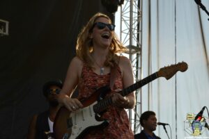 Female guitarist on stage, smiling while playing a white Fender Stratocaster, wearing sunglasses and a patterned dress.