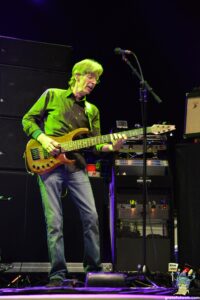 Bass guitarist on stage under green lighting, playing a wooden bass with a strap, microphone stand nearby.