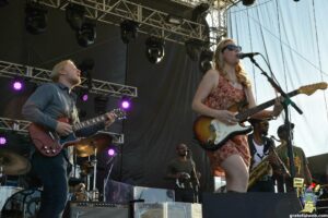 Female vocalist wearing sunglasses sings into a microphone while playing an electric guitar on a stage, with a guitarist beside her and stage rigging overhead during a live outdoor concert.