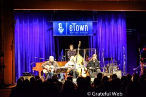 Live concert scene with a band playing guitars and singing on a stage framed by blue curtains and a banner that reads "eTown"; audience seated in front.