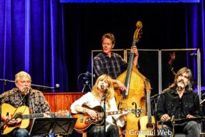 Live band on stage playing guitars and singing, blue curtain backdrop, angles of string instruments visible