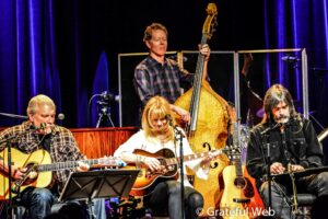 A jazz/folk band performing on stage: musicians with guitars, a bassist at center, blue-curtain backdrop, audience-facing scene.
