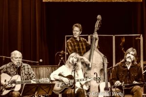 Live band on stage performing a song, with guitarists and a upright bass visible in a sepia-toned scene.