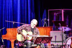 Older man in a plaid shirt playing an acoustic guitar on a stage with blue curtains in the background
