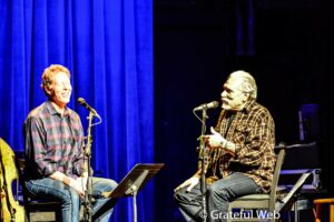 Two male singers in plaid shirts seated on stage, singing into microphones with a blue curtain background.