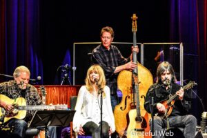 Female vocalist seated front, surrounded by male guitarists and a bassist on stage during a live band performance.