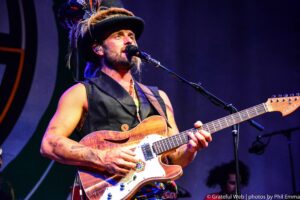 Male guitarist singing into a microphone while playing a sunburst electric guitar, wearing a feathered hat under blue stage lighting.