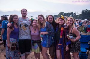 Group of six friends posing together at an outdoor festival, smiling, with colorful boho outfits and drinks in hand.