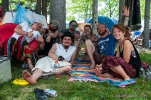Group of seven friends posing for a photo at a forest campsite, sitting on colorful blankets with tents in the background and trees overhead, looking relaxed and happy.