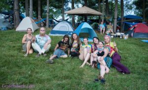 Group of friends with colorful ties and t-shirts sitting on grass at a forest campground, tents in the background, smiling at camera and flashing peace signs.