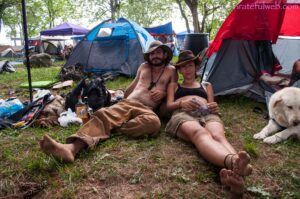 Couple in hats lounging on grass at a busy campsite with tents and a dog nearby.