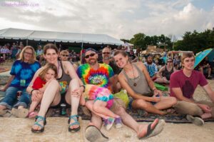 Smiling group seated on a blanket at an outdoor festival with tents and many people in the background.
