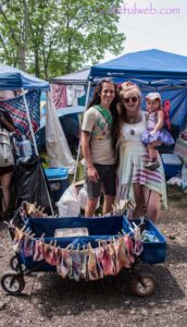 Family of three posing at an outdoor festival or market booth; a wagon of colorful socks hangs on a clothesline in the foreground with blue tents in the background