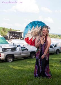 Smiling woman in a colorful patterned dress holds a blue umbrella parasol at an outdoor festival, with a stage and parked trucks in the background.
