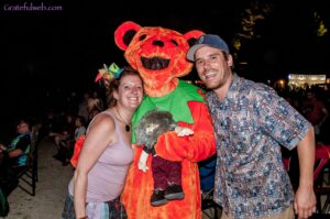 Two adults smiling with a large orange bear mascot at a nighttime outdoor event.