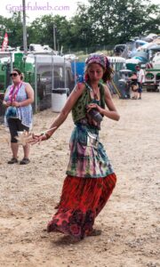 Young woman in a colorful bohemian outfit dancing on a gravel festival ground, with tents and people in the background.