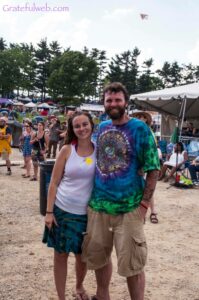 Smiling couple at an outdoor festival with tents and a crowd in the background.