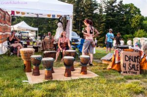 Outdoor festival scene with a row of carved djembé drums and people sitting and chatting under tents near a grassy area.