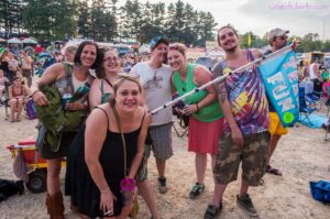 Group of friends posing for a selfie at a large outdoor festival, tents and trees in the background (sunny day).