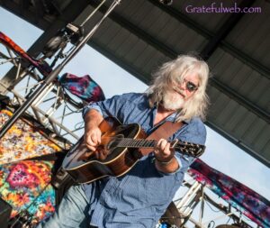 Musician with long gray hair and sunglasses playing an acoustic guitar on an outdoor stage, with colorful banners in the background
