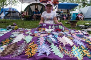 Smiling woman with colorful hair sits behind a purple blanket covered with handmade patches at a campsite with tents in the background.