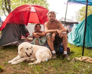 Two campers sit near a red tent at a sunny campsite, with a large white dog resting on the grass in front of them.